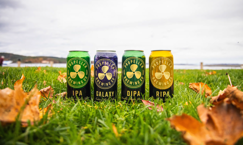 Photo of four cans of Propeller beer, lined up in the grass with a gray sky behind them. From left to right, the cans of beer are IPA, Galaxy DIPA, DIPA and RIPA (Rye IPA)