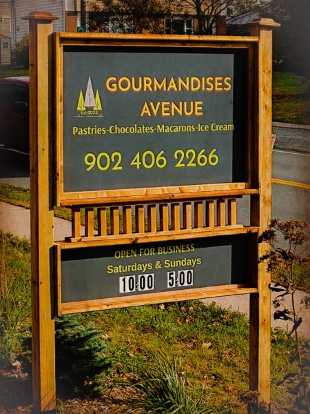Sign for Gourmandises chocolate shop. Wooden trim with grey background on front lawn of their store. 