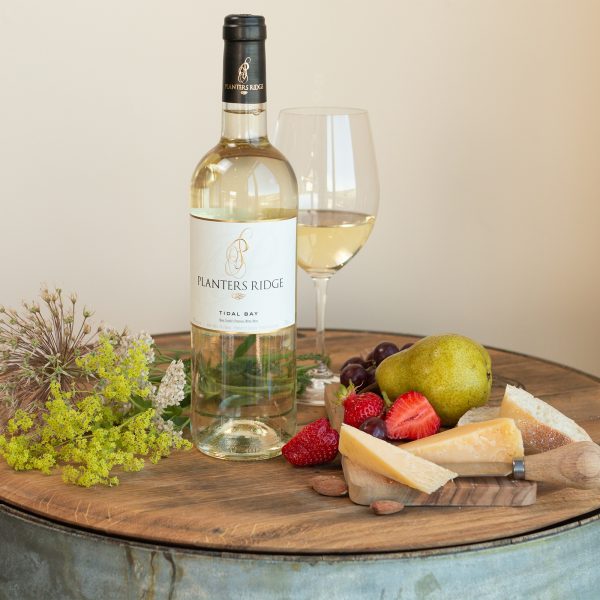 A Tidal Bay wine bottle sitting next to a wine glass, various fruit and cheese. 