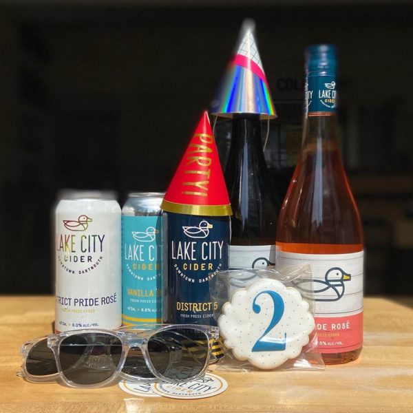 Photo of Lake City Cider's 2nd birthday bundle, featuring two bottles of cider (one wearing a hat), three cans of cider (one wearing a hat), two pairs of sunglasses and a round cookie that has the number 2 written on it.