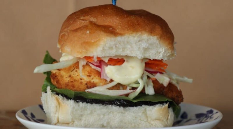 Photo of a halibut burger on a white patterned plate. The halibut is on a white hamburger bun and toppings include tarter sauce, lettuce, slaw, carrots, radishes and a blueberry sauce.