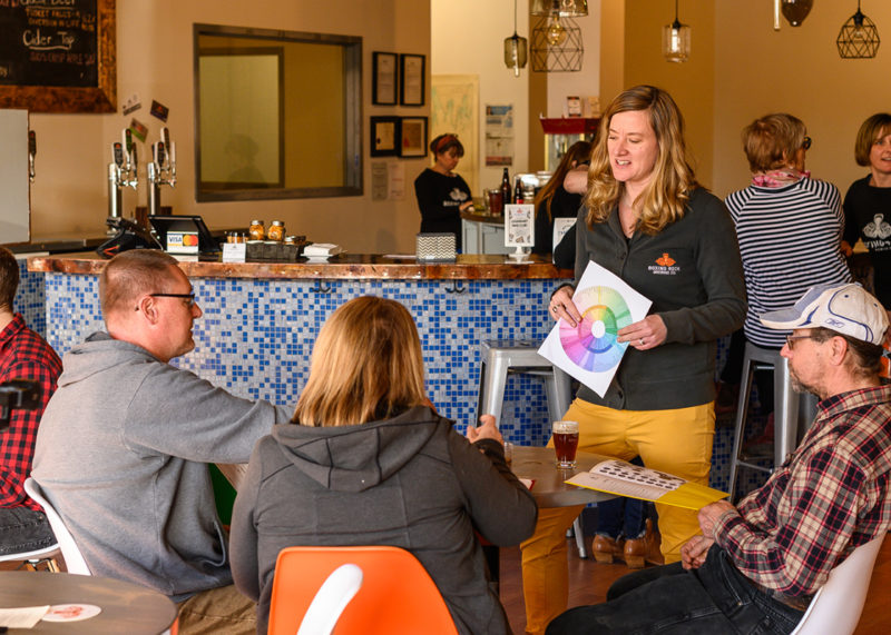 A photo of the Craft Beer 101 experience at Boxing Rock Brewery. In the photo, a woman wearing a Boxing Rock shirt is standing in front of a table of three guests (two men, one female) and holding a sheet, while talking to them about beer.