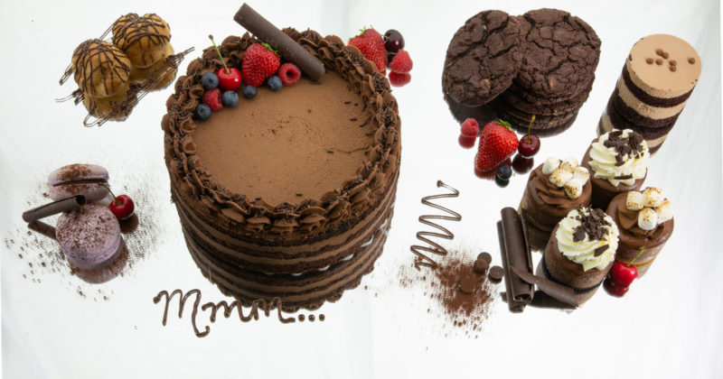 A photo of the various products Delectable Desserts makes, including chocolate cake with berries on top, chocolate macarons, chocolate cookies, mini cheesecakes and more.