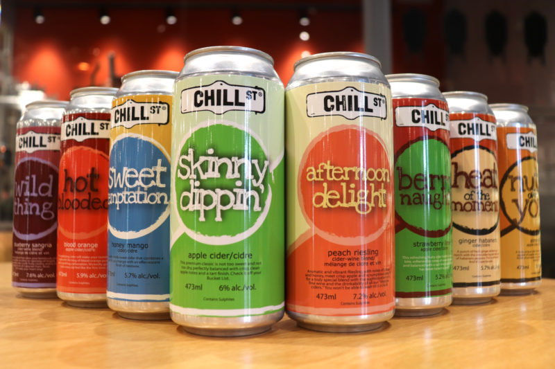 A photo of 8 different cans of Chill Street craft cider products. From left to right: A can of Wild Thing (Blueberry Sangria), Hot Blooded (Blood Orange), Sweet Temptation (Honey Mango), Skinny Dippin' (Apple Cider), Afternoon Delight (Peach Reisling), Berry Naughty (Strawberry Lime), Heat of the Moment (Ginger Habanero), Nuts Over You (Almond)