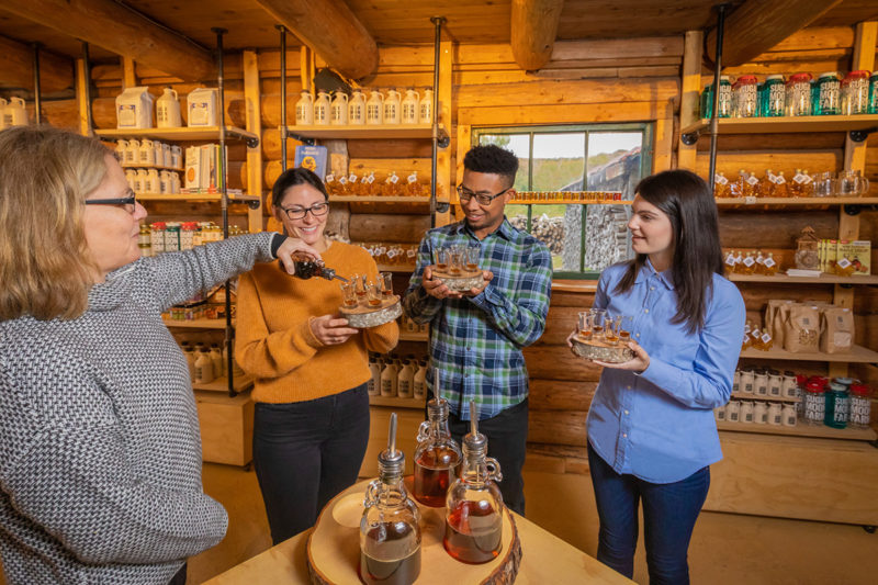 A photo of the Canadian Maple Indulgence package at Sugar Moon Farm. Photo shows three people standing around holding small flights of maple syrup. Owner Quita Gray (left hand side of photo) is pouring maple syrup into a small container.