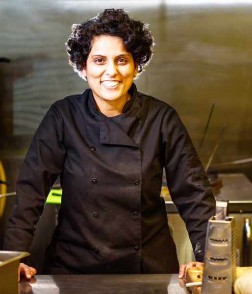 Photo of Shivani Dhamija, owner of Shivani's Kitchen, standing alone in a kitchen. She is smiling and wearing a black chef's jacket.