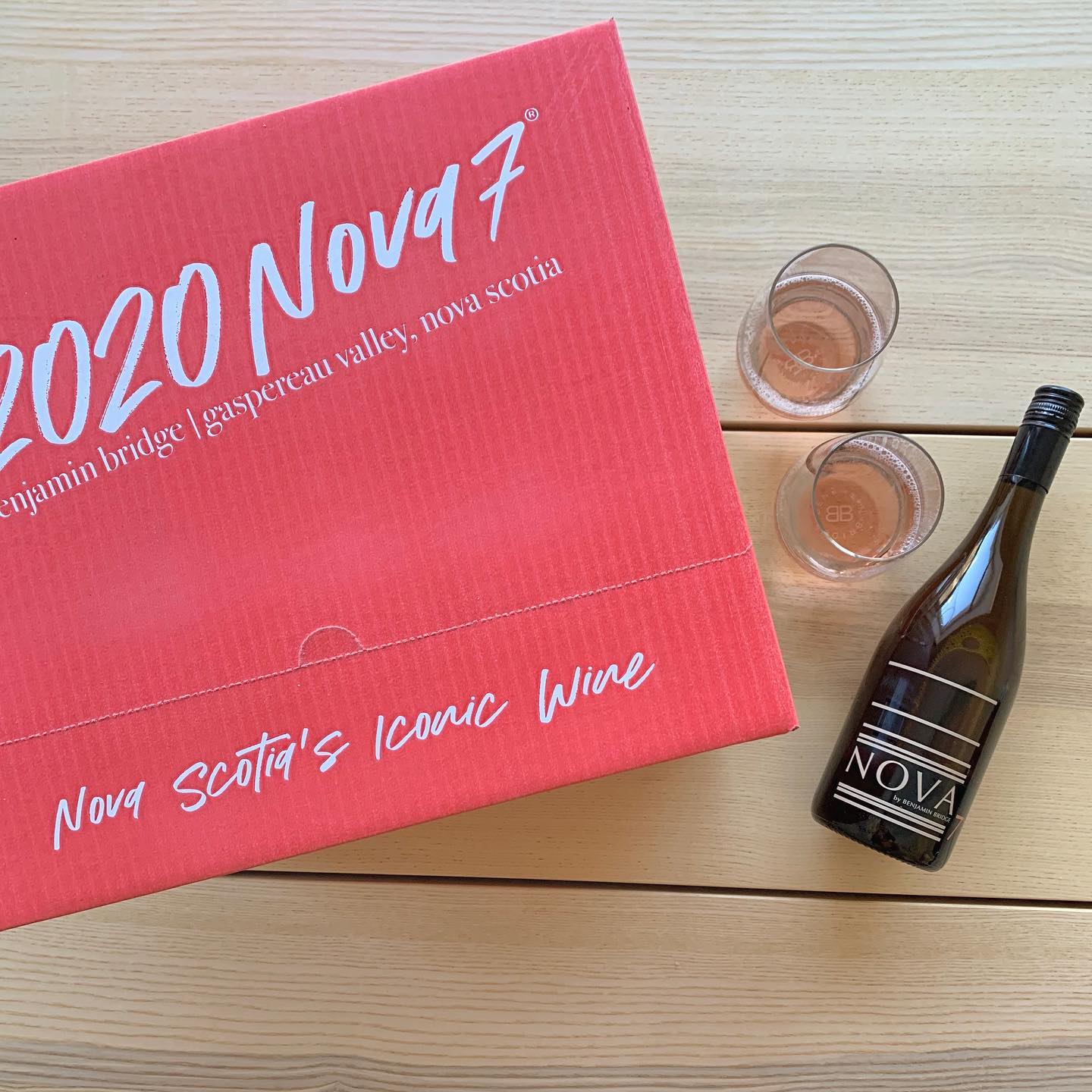 Image of a bottle of Nova 7 with 2 filled wine glasses and pink shipping box