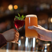 Two people cheers at a bar. One is holding a red cocktail with a mint garnish and the other holds an amber coloured beer.