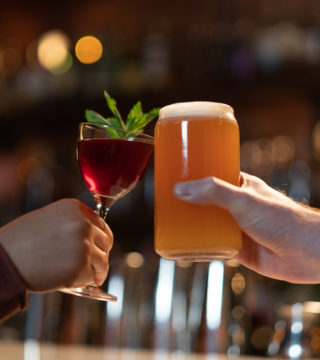 Two people cheers at a bar. One is holding a red cocktail with a mint garnish and the other holds an amber coloured beer.