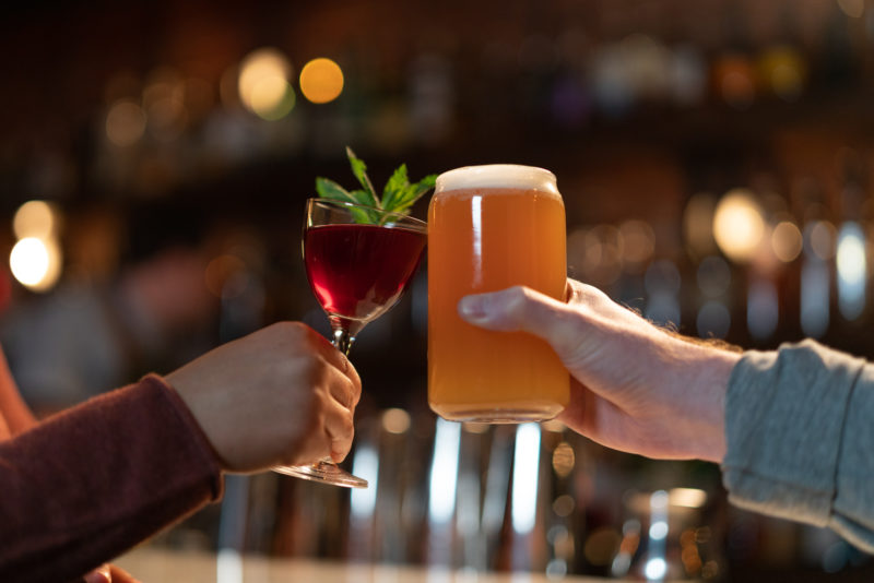 Two people cheers at a bar. One is holding a red cocktail with a mint garnish and the other holds an amber coloured beer.