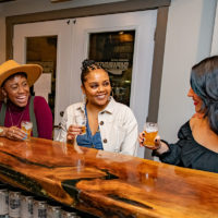 Three women smile and enjoy beer from Shipwright Brewing Company at the wooden bar