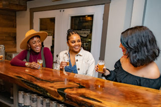 Three women smile and enjoy beer from Shipwright Brewing Company at the wooden bar