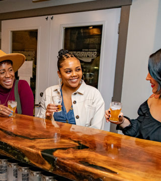 Three women smile and enjoy beer from Shipwright Brewing Company at the wooden bar