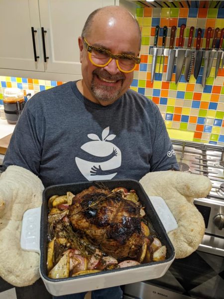 The kilted chef holds a roasted pork with apple and parsnips. He is wearing beige oven mats and yellow glasses. 