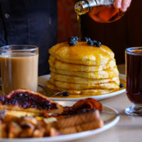 Person pours maple syrup over a stack of pancakes. Coffee, tea, toast and hash browns are in the foreground.