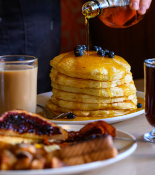 Person pours maple syrup over a stack of pancakes. Coffee, tea, toast and hash browns are in the foreground.