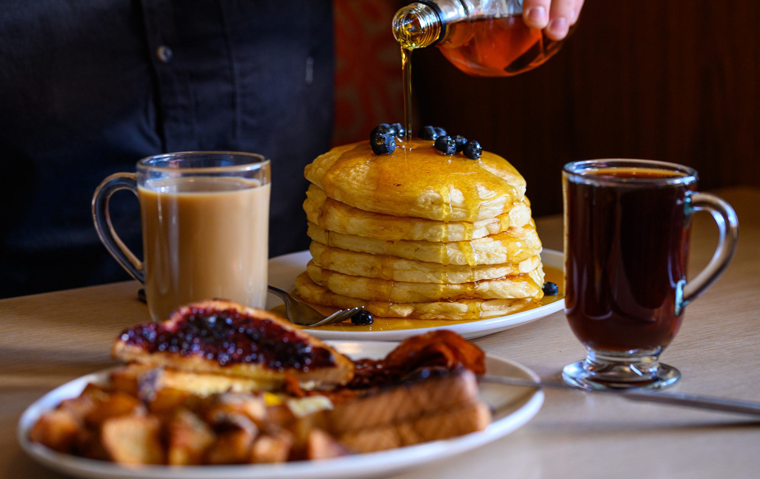 Person pours maple syrup over a stack of pancakes. Coffee, tea, toast and hash browns are in the foreground.