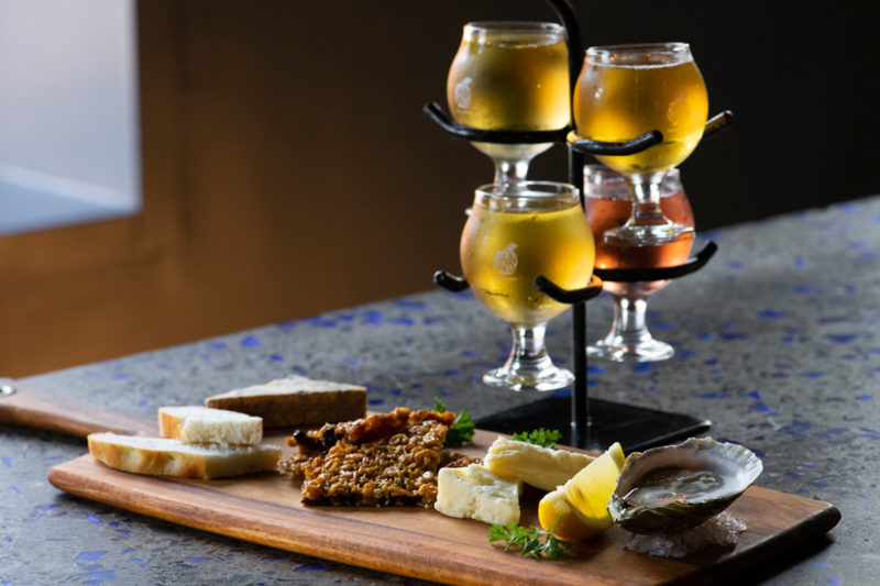 A photo of a flight of four different ciders, paired with a small snackboard with breads, cheese, oysters and more. The photo is a part of the Pommelier experience at Chain Yard Urban Cidery.