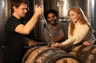 A photo of three people standing around a barrel in a distillery - one man (the head distiller) has drawn liquour from the barrel and is pouring it into the glass of a woman in a white sweater. There is a man standing beside her, holding a glass and looking at the liquor being poured.