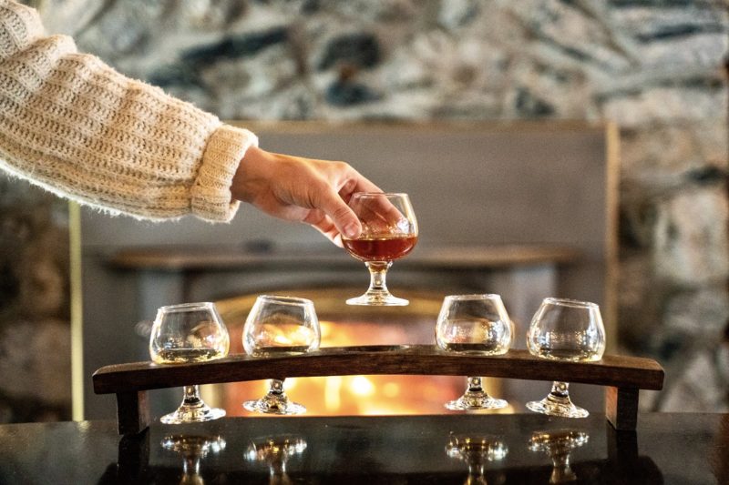 Woman holds up a whisky tasting glass in front of a fire. There are four other tasting glasses in the image. There is a stone wall in the background.