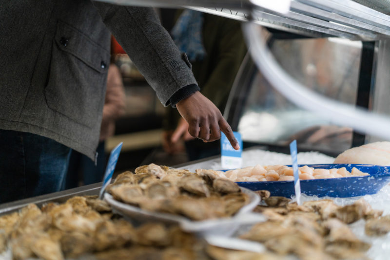 Man points to scallops inside seafood counter. There are also oysters in the shot. 