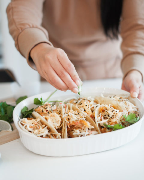 Woman places piece of cilantro on fish tacos. Tacos are in a white platter and the woman is wearing a tan coloured long sleeve shirt. 