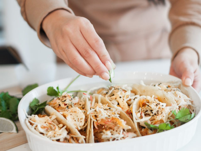 Woman places piece of cilantro on fish tacos. Tacos are in a white platter and the woman is wearing a tan coloured long sleeve shirt.