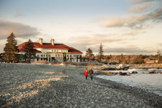 Une photo d’un homme et d’une femme marchant sur la plage rocheuse devant le White Point Beach Resort. Le pavillon principal est visible à l’arrière-plan ; l’homme porte un manteau noir tandis que la femme porte un manteau rouge et une écharpe.