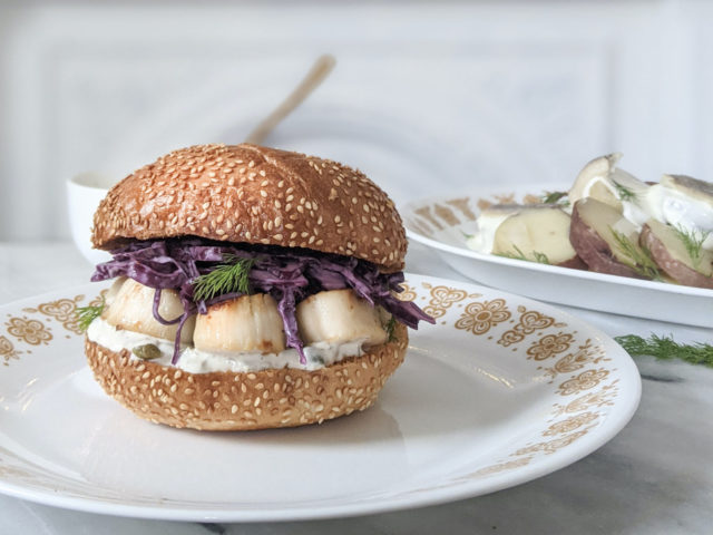 scallop burger on sesame bun sitting on antique white plate with yellow detailing