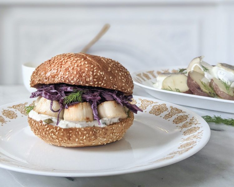 scallop burger on sesame bun sitting on antique white plate with yellow detailing