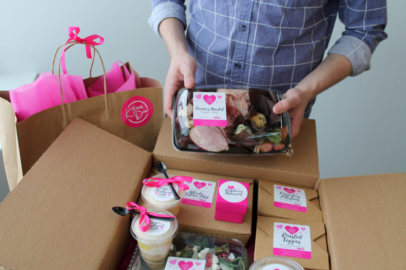 Person in a denim shirt opens a cardboard box filled with small boxes of food from Kitchen Door. 