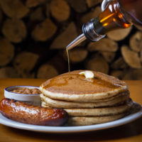 pancakes, sausage and baked beans sit on a plate in front of stacks of wood. Maple syrup is poured on them.