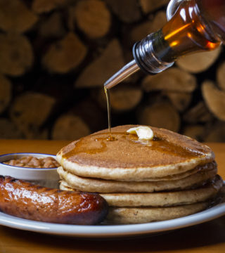 pancakes, sausage and baked beans sit on a plate in front of stacks of wood. Maple syrup is poured on them.