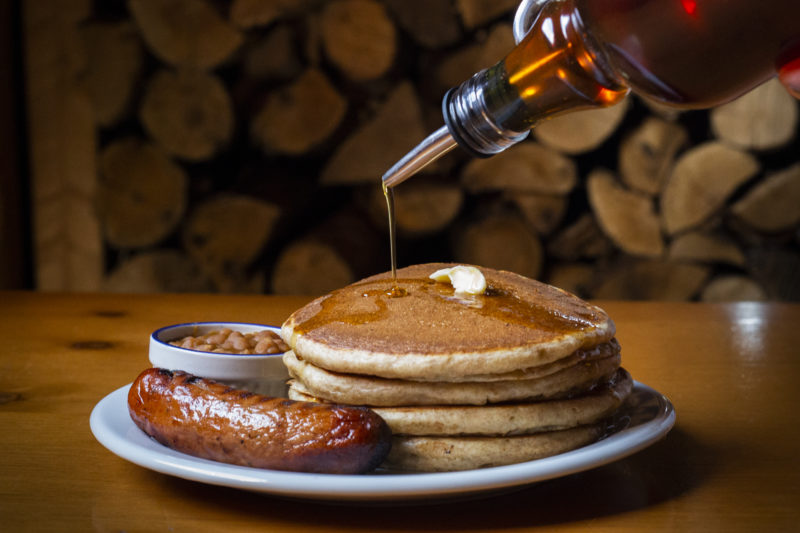 pancakes, sausage and baked beans sit on a plate in front of stacks of wood. Maple syrup is poured on them.