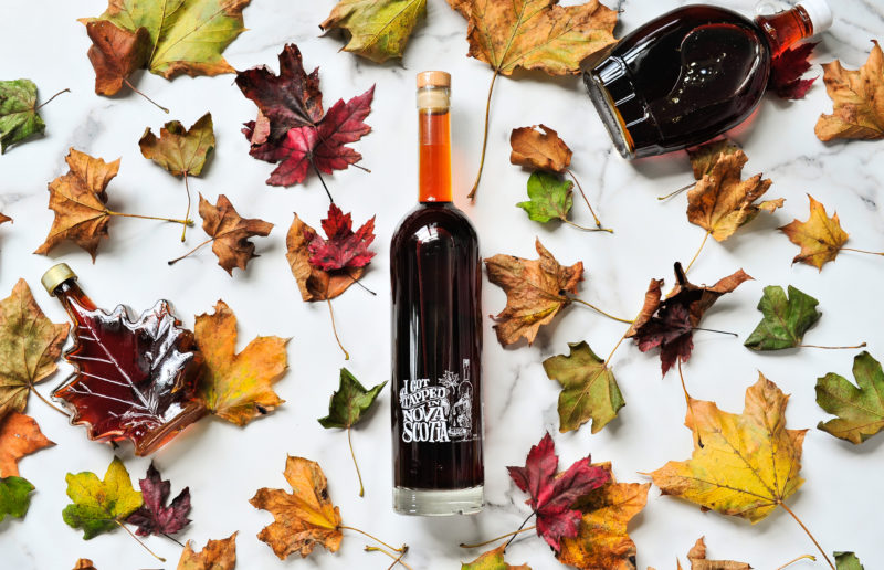 Bottle of Steinhart Distillery vodka sits in the middle of image. Maple leaves are scattered throughout the image with maple syrup bottles to the left and right. 
