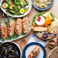 Salad, lobster rolls, mussels, potato salad, and assorted cheese laid out on various sized plates on wooden table.