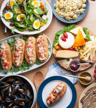 Salad, lobster rolls, mussels, potato salad, and assorted cheese laid out on various sized plates on wooden table.