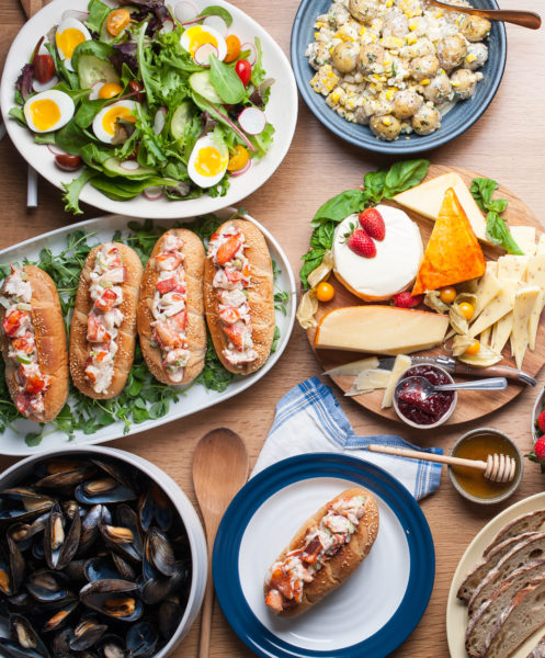 Salad, lobster rolls, mussels, potato salad, and assorted cheese laid out on various sized plates on wooden table.