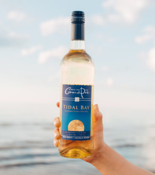 a hand holds a bottle of Grand Pre Tidal Bay in front of the ocean