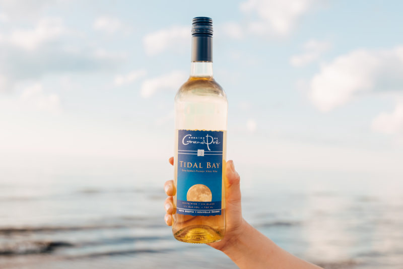 a hand holds a bottle of Grand Pre Tidal Bay in front of the ocean