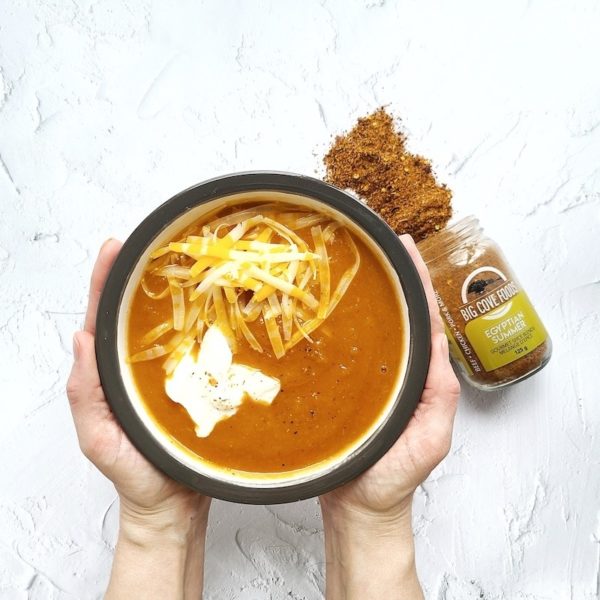 two hands hold a bowl of soup with cheese and sour cream. A big cove food spice jar is open and spices are displayed next to the bowl.