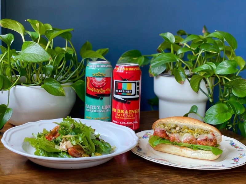 A salad and a meatball sub sit on white plates with two beer in the background centre and two potted green plants on each side. 