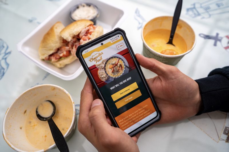 Hands hold a phone with the Nova Scotia Chowder Trail website on it. Two bowls of chowder and a lobster roll are also in the photo