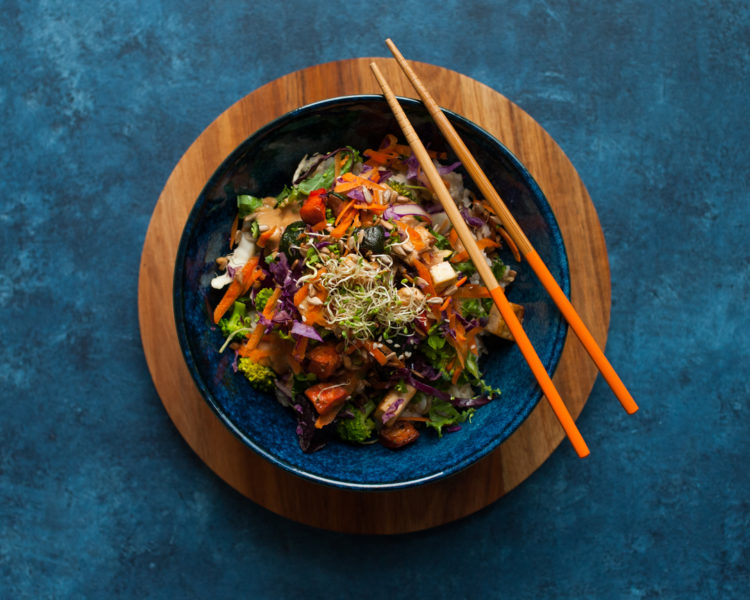 blue bowl filled with a vegetable dish sits on a wooden platter on top of a blue background. two wooden chopsticks are on the bowl. 