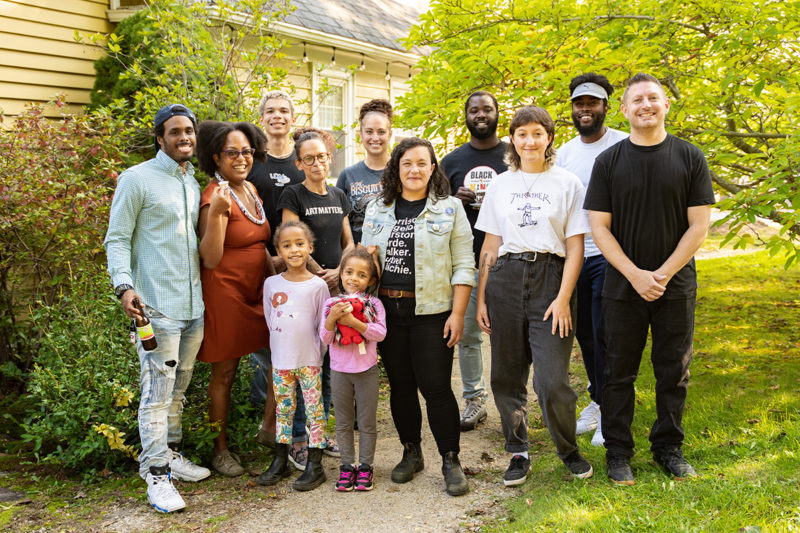 A group of people stand outside a building in front of green trees and green grass. 