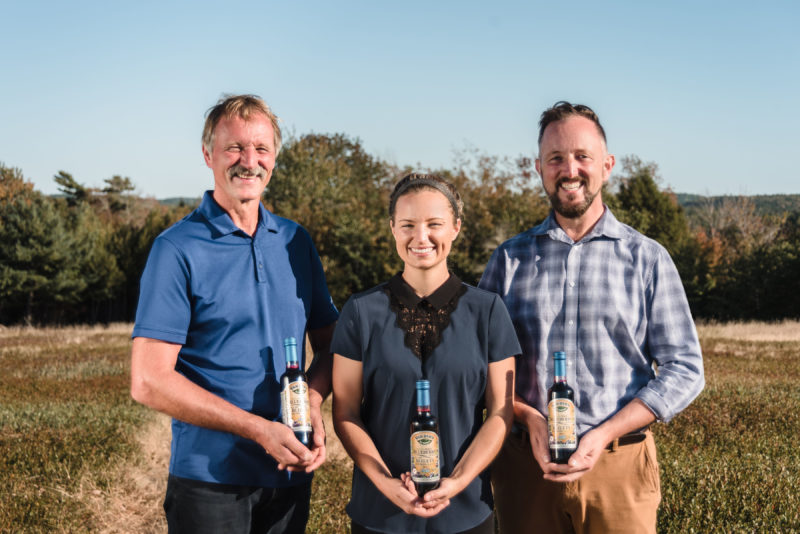 Three people stand in a blueberry field holding bottles of blueberry juice