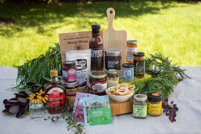 several local products in jars and packages sit on top a cutting board. A smaller cutting board is in the background. There is lots of greenery around the items. 