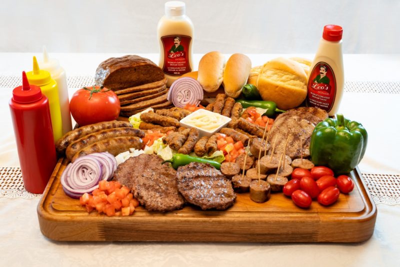 A photo of a spread of food on a wooden cutting board to help promote Leo's Donair. The donair burgers, sausages, loaves and donair sauce are all from Leo's Donair.