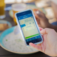 A hand holds a phone with the Taste of Nova Scotia app open over a bowl of chowder