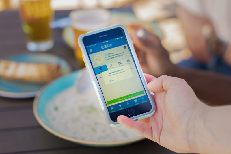 A hand holds a phone with the Taste of Nova Scotia app open over a bowl of chowder